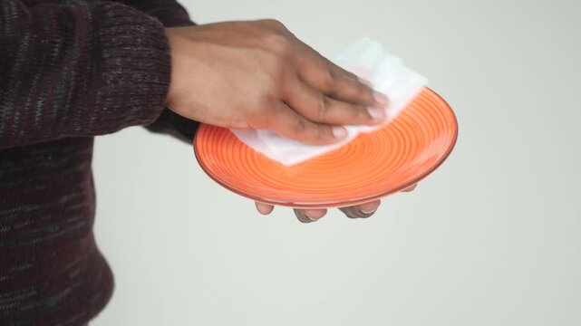 Person cleaning orange ceramic plate with white tissue paper against light background