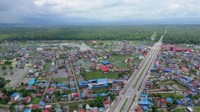 Elevated aerial perspective of a long straight multi-lane highway cutting through a vibrant urban settlement toward a dense green forest in Nepal.