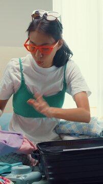Vertical Screen : Young happy and exciting Asian female ready for summer holidays prepare suitcase put cloth on her bag in full of messy stuff bedroom. Preparing luggage for summer vacation trip.