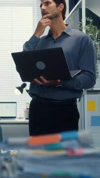 Vertical Screen : Thoughtful young Latin businessman stand with laptop analyzing data paperwork stack. Hybrid work, digital transformation strategy, business corporate finance, executive decision.