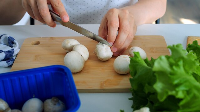 Person prepares raw champignon mushrooms on wooden board with metal kitchen knife, creating potential kitchen injury risk as bare fingers stabilize vegetable dangerously near sharp steel blade.