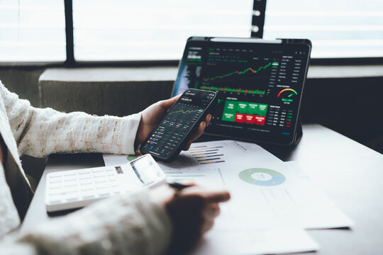 Investor using a smartphone to monitor stock market data while analyzing financial charts and investment reports on a laptop.