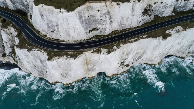 Top down aerial view of asphalt road winding along edge of steep white chalk cliffs above turquoise ocean 