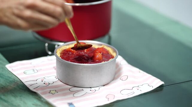Woman layering the mixture of paneer, curd and honey on the layer of crushed biscuits in a bowl, and adding the strawberry puree over it - in process of making strawberry cheesecake, kitchen.