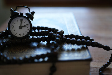 The Quran, clock, and prayer beads on a wooden table with dim lighting. Creates an atmosphere of spiritual contemplation and a sense of reverence. Reminds us of the time to pray. Muslim, Ramadan, Isla © PANCASONA CREATION