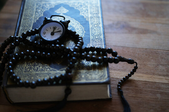 A Quran, a clock, and prayer beads on a wooden table evoke a moment of spiritual reflection and reverence. A background image suitable for Muslims, Eid al-Fitr, Eid al-Adha, and the Muharram New Year.
