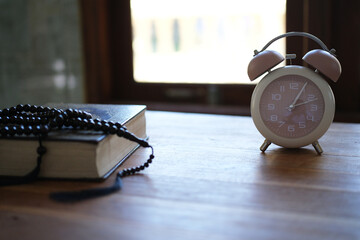 The Quran, clock, and prayer beads on a wooden table with dim lighting. Creates an atmosphere of spiritual contemplation and a sense of reverence. Reminds us of the time to pray. Muslim, Ramadan, Isla © PANCASONA CREATION