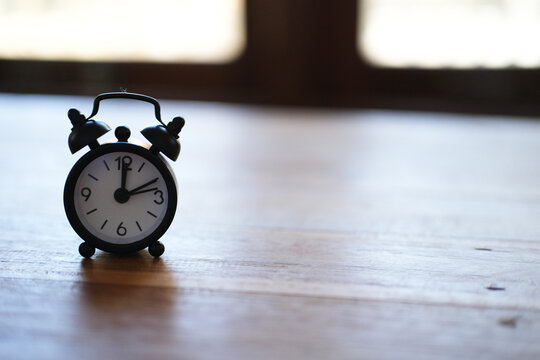 An alarm clock sits on a wooden table near a window, silhouetted against the light. It represents reminders and time management. The scene features an empty table and a stylish alarm clock&mdash;a classic s