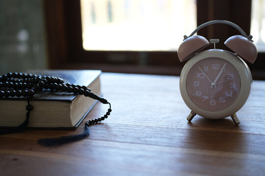 The Quran, clock, and prayer beads on a wooden table with dim lighting. Creates an atmosphere of spiritual contemplation and a sense of reverence. Reminds us of the time to pray. Muslim, Ramadan, Isla