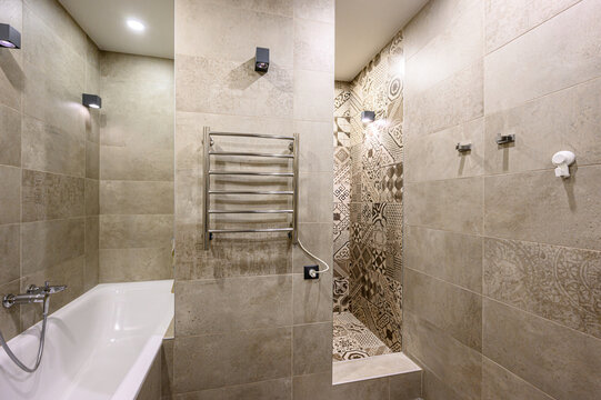 Compact bathroom with beige stone tiles, geometric pattern accent wall, built-in bathtub, chrome towel warmer, recessed lighting, wall sconces, shower hose, wall fixtures