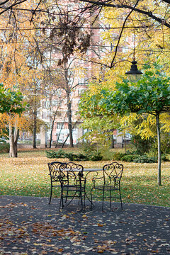 Elegant autumn park with wrought iron garden chairs and table on a leafy terrace, surrounded by golden trees, fallen leaves and vintage street lamp, creating a peaceful seasonal atmosphere