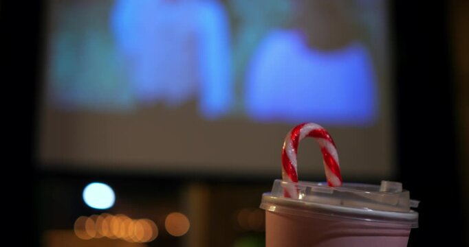 Striped candy cane sticks out from disposable beverage cup in home cinema. Festive sweet drink provides cozy refreshment while projector illuminates blurred background with warm holiday lighting