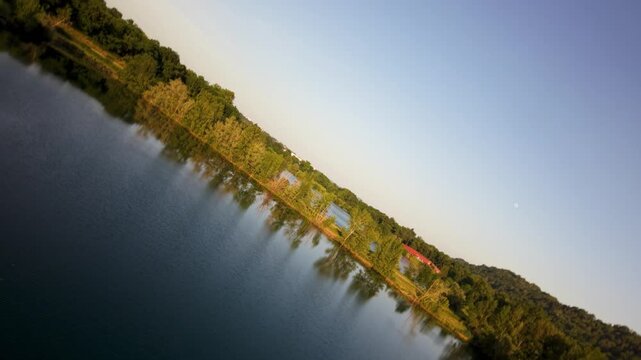Aerial spin roll 180 shots show a lake with trees lining the shore and buildings visible in the background. The surroundings are green
