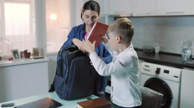 Dark-haired parent helping her kid pack a school backpack indoors.