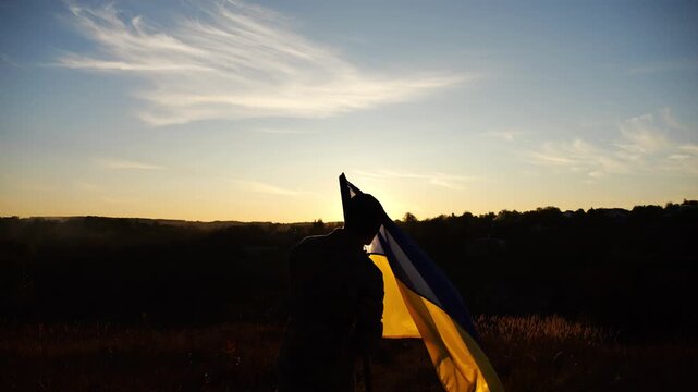 Silhouette of military man kissing blue-yellow flag and raising it up at countryside. Male ukrainian army soldier with lifted national banner in honor of victory against russian aggression