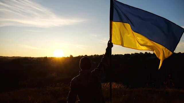 Male military in uniform lifting blue-yellow banner in honor of the victory against russian aggression at sunset. Young soldier of ukrainian army raised a waving flag of Ukraine at countryside