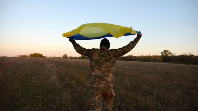 Soldier of ukrainian army running with raised flag Ukraine above his head at field. Young male military in uniform jogging with blue-yellow banner at meadow. Victory against russian aggression