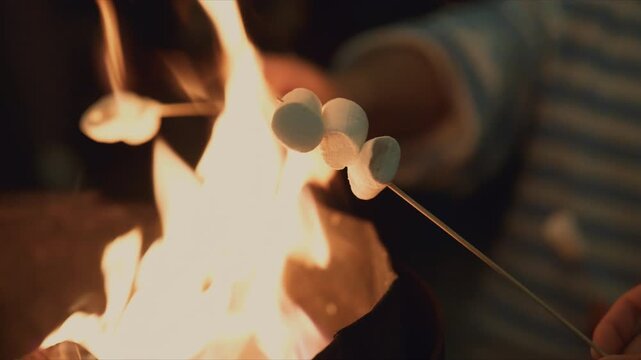 Close up of hands roasting marshmallows on sticks over a campfire at night