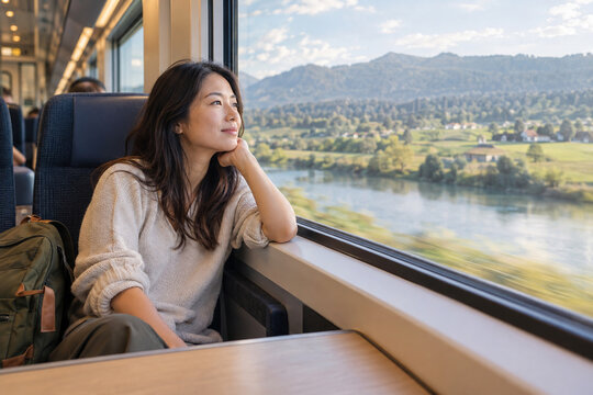 Serene Journey: A woman gazes out the train window at a picturesque landscape of rolling hills, a tranquil river, and lush greenery, embodying a moment of peaceful contemplation and travel.