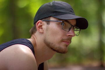 Side profile portrait of a thoughtful young man with glasses, stubble, and a black mesh cap sitting in a summer forest © nc_lia
