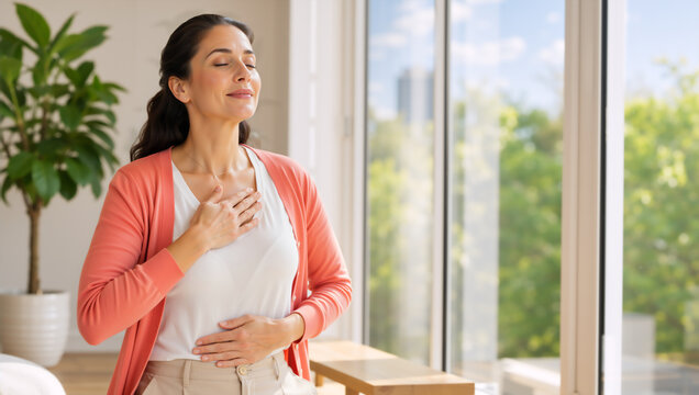 Woman practicing deep breathing with hands on chest and stomach. Mindful female meditating near window at home. Wellness and self-care concept
