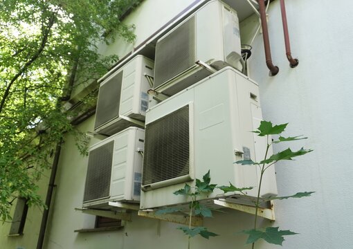 Air conditioning units on building exterior. Multiple white AC units mounted on an exterior wall with green trees in the background. 