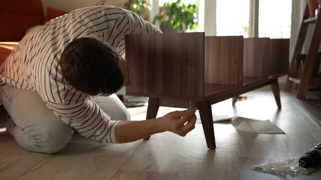 Concentrated man assembling new furniture for his apartment, kneeling on the floor and carefully tightening screws on a wooden tv stand, enjoying a diy home improvement project