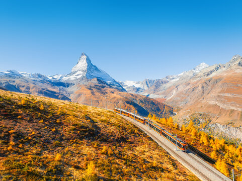 Aerial drone view of a scenic railway line curving through a colorful autumn forest with Matterhorn peak in Zermatt, Switzerland. Bright sunny day alpine landscape.