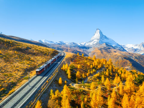 Aerial drone view of a scenic railway line curving through a colorful autumn forest with Matterhorn peak in Zermatt, Switzerland. Bright sunny day alpine landscape.