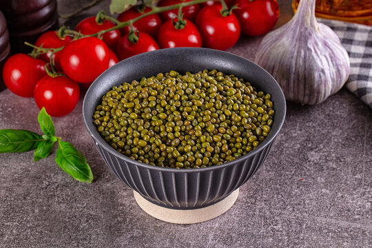 Green mung beans in textured black bowl with cherry tomatoes, garlic bulb and basil leaves on gray stone surface