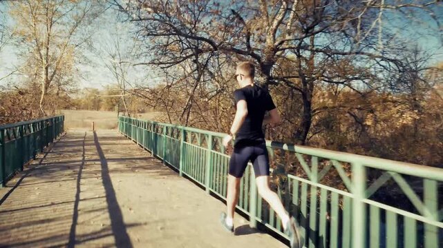 Runner Jogging Across Wooden Bridge Over River In Autumn Park. Outdoor Training. Exercise Cardio Workout
