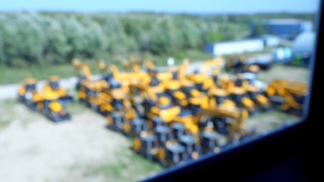 Blurred view of many yellow construction vehicles parked outdoors. Background shows green trees and open sky. Image appears through a window frame. Scene remains out of focus across all frames