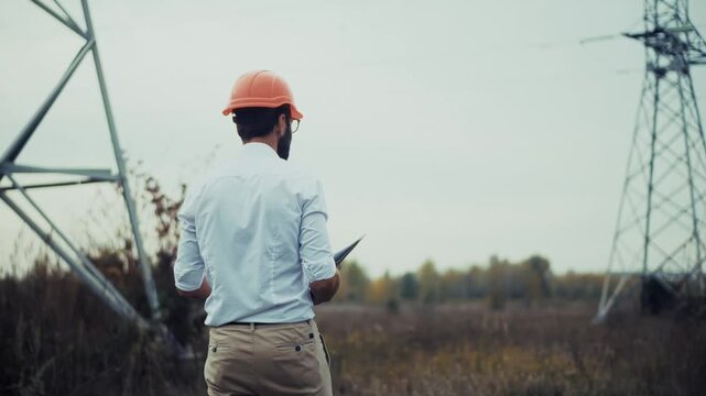 Electrical Engineer In Hardhat Holding Tablet Inspecting High Voltage Power Line Pylon. Electricity Energy Grid Substation Maintenance 