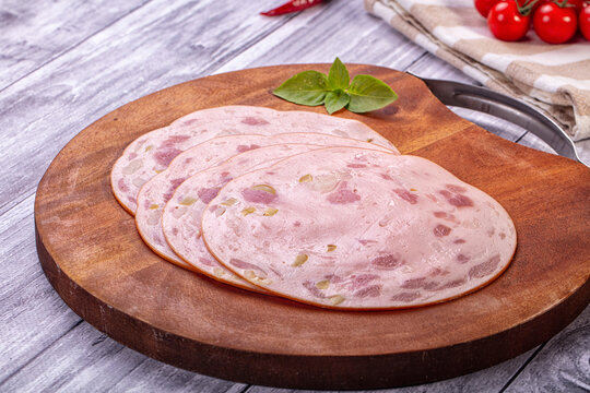 Sliced mortadella with visible olive pieces on wooden cutting board, fresh basil leaf garnish, cherry tomatoes and checkered cloth background