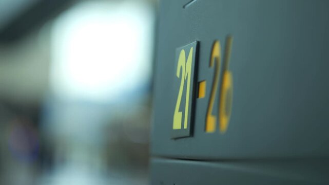 Close-up of airport gate signs showing numbers 21-26. Blurred background with bright lights and indistinct shapes. Focus on metallic surface with yellow lettering