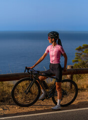 A full-length profile portrait of an athletic female cyclist wearing a helmet and a pink jersey, standing astride her road bike on a coastal road and happily gazing out at the Mediterranean Sea. © sirbouman