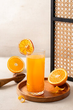 Fresh orange juice in a tall glass with dried orange slice garnish on a wooden tray, styled with halved oranges, wooden citrus juicer, and rattan frame