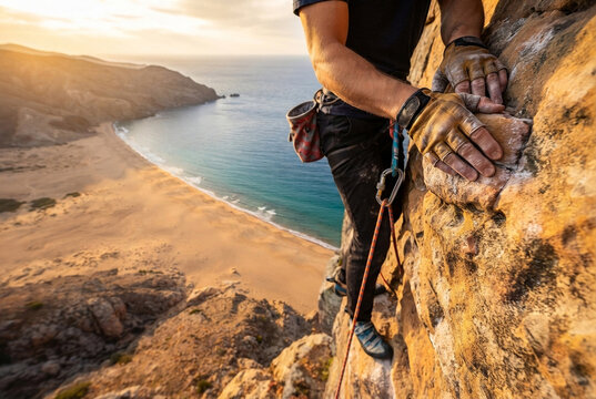 A rock climber ascends a steep cliff overlooking a scenic beach.