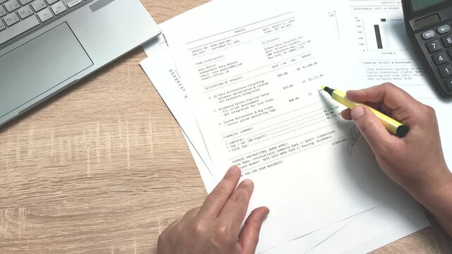 Top view overhead shot of contractor hands checking printed invoice document on wooden desk and highlighting total amount in yellow pen, preparing payment paper in office.