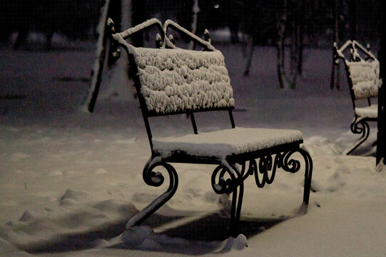 A city park on a winter evening. A snow-covered bench in the light of a streetlamp.