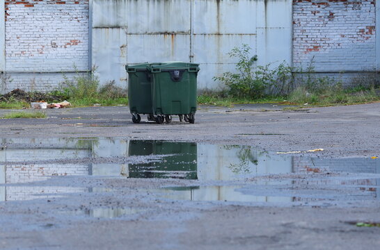 A lone green plastic trash bin in an empty courtyard with puddles on the asphalt