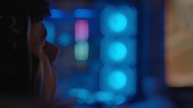 Close-up of a young gamer concentrating on a video game, his face illuminated by the vibrant, changing colors of his RGB-lit computer setup in a dark room during an intense online match