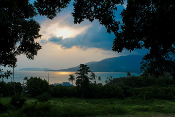Dramatic tropical sunset over the seaside, with golden sun rays breaking through layers of clouds and illuminating the sky above the calm ocean. The warm evening light creates a striking atmosphere, w © Yuliia