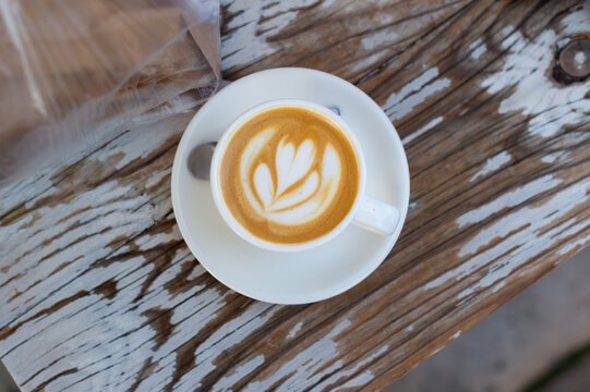 A top-down view shows a cup of cappuccino with latte art served on a white saucer. The cup sits on a rustic wooden table outdoors in Kerkira, Greece.
