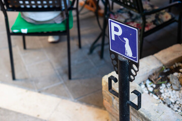 A dog parking sign on a metal post stands outside a cafe in Kerkira, Greece. The blue sign features a dog silhouette and a leash attachment hook below. © dsbedarieva