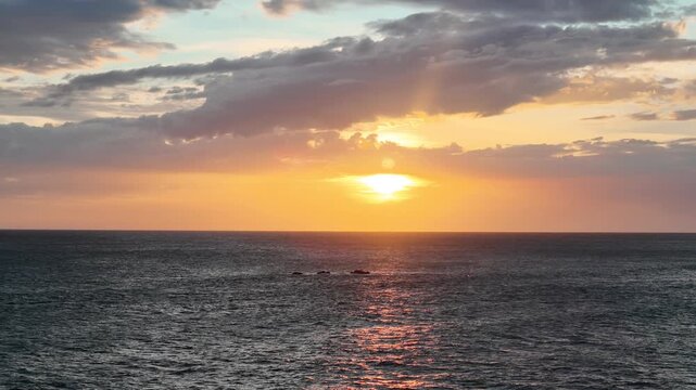 The sun sets over the ocean casting warm colors across the water. Clouds float in the sky as rocks are visible on the horizon.