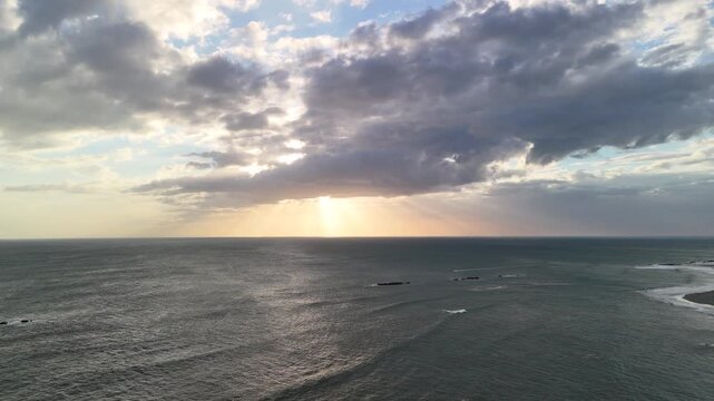 Waves roll in as the sun sets on the horizon. Clouds scatter across the sky. The scene captures the beauty of the ocean.