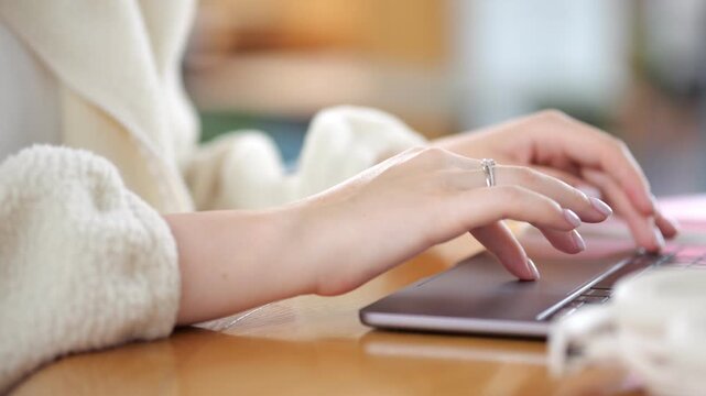 Close-up of hands typing on laptop keyboard. Fingers move quickly over keys. Ring visible on left hand. Soft focus background shows cozy indoor setting. Warm wooden table surface beneath device