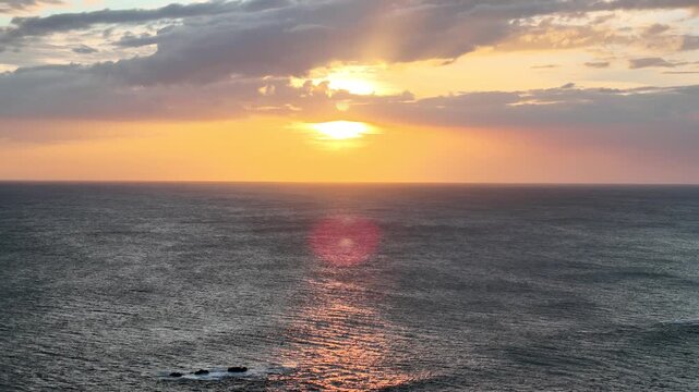 A sunset is seen over the ocean. The sky has clouds and colors as the sun sets. Waves gently move in the water.