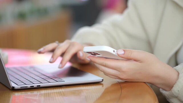 Person uses phone and laptop on wooden table. Hands interact with devices closely. Background shows blurred indoor space